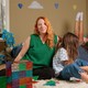 A woman sits on the floor of a bedroom while three children play by themselves.