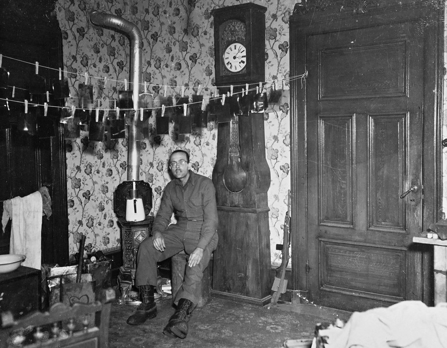 A U.S. Army soldier sits in a room in a house in France, beneath several lines where many film negatives hang to dry.