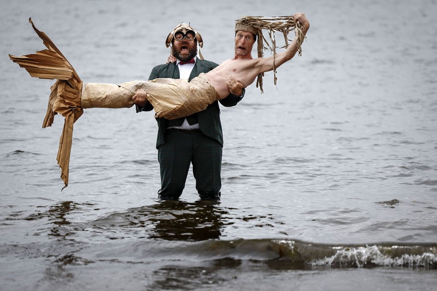 Two performers pose for a photo in shallow water, one holding the other (dressed as a mermaid) in their arms.