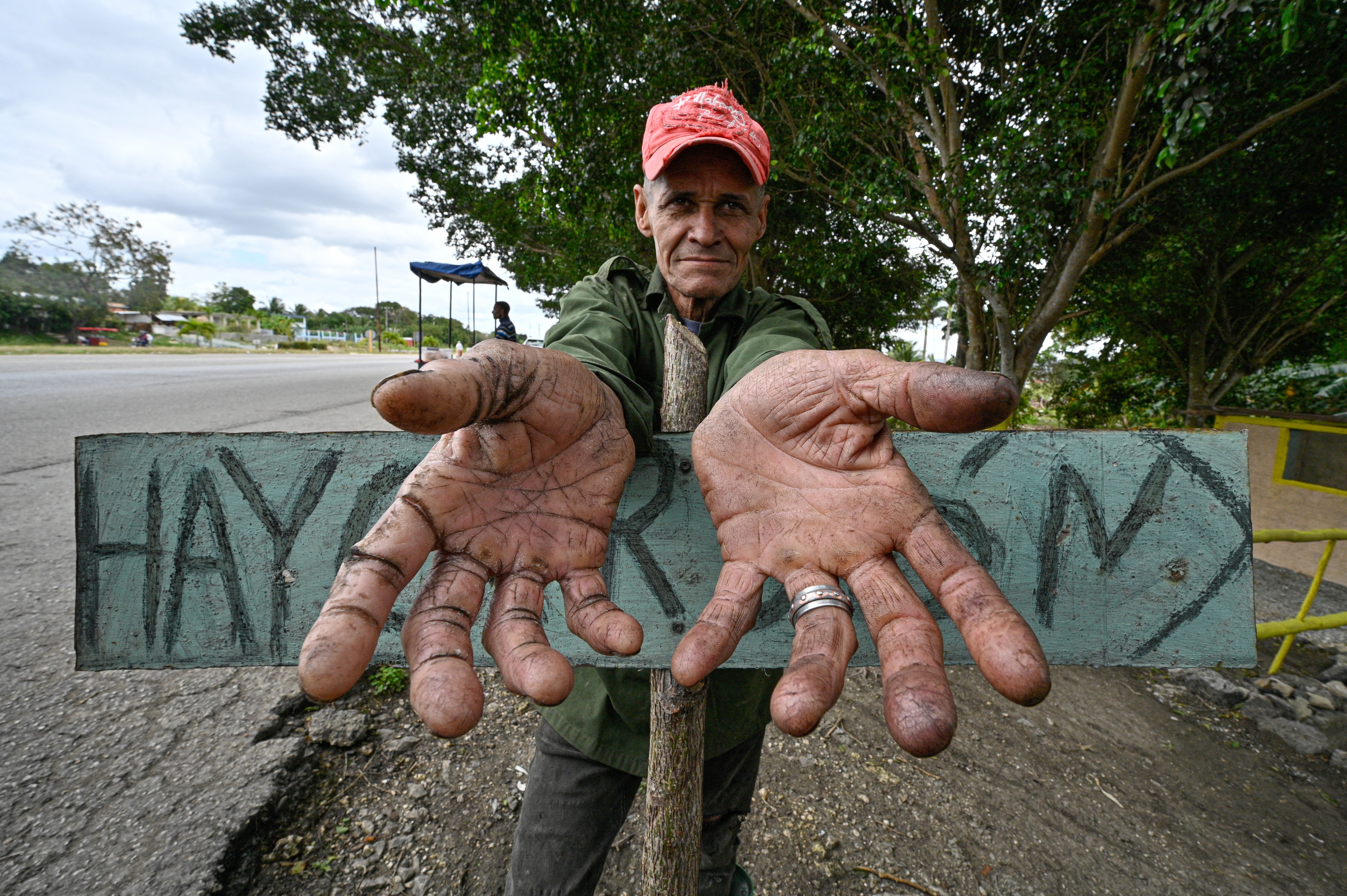 A charcoal seller shows his dirty hands as he waits for customers, leaning on a small sign.