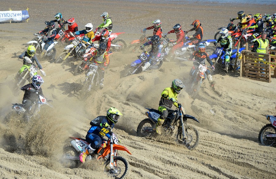 More than a dozen motorcycle riders make a turn during a race on a sandy beach.