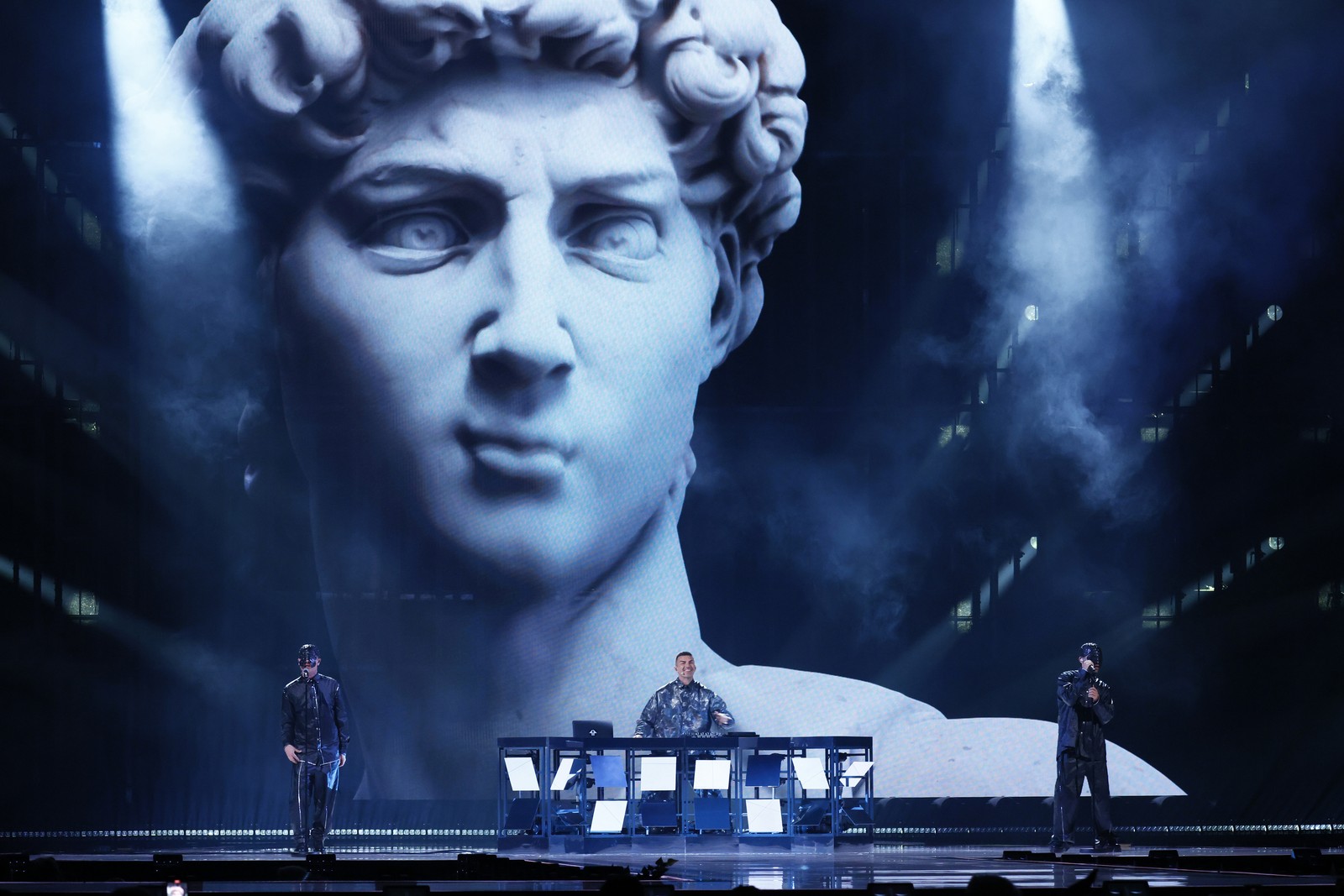 A DJ and singers perform in front of a large video screen, showing an animated Statue of David.
