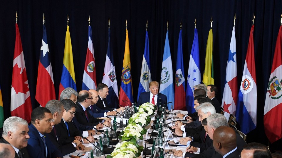 President Donald Trump sits at the head of a table surrounded by leaders from other countries and their flags.