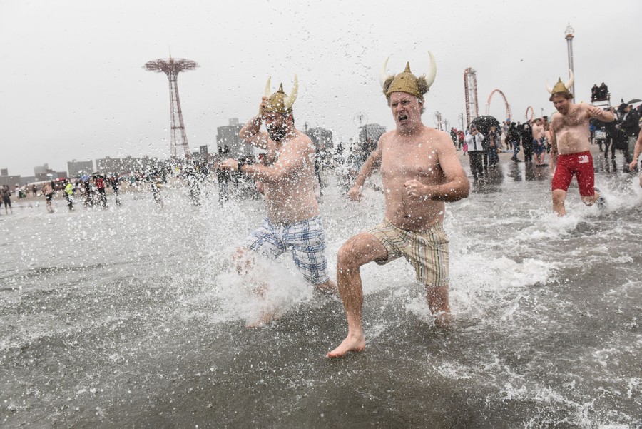 Several people in bathing suits and costume hats are seen running into cold surf.
