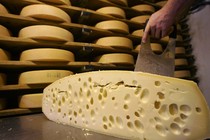 A hand slices cheese in front of shelves of more cheese.