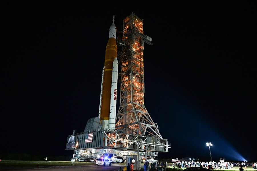 A tall rocket ship stands on a mobile platform beside a tall tower, lit up at night.