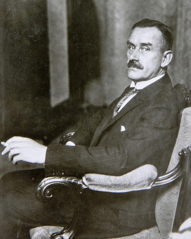 A black and white photograph of Thomas Mann sitting in a suit looking over at the camera on his left
