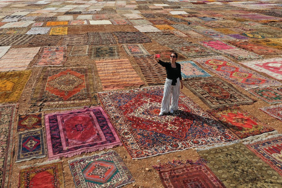 A person takes a selfie in a field covered in handmade rugs.