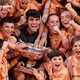 A tennis player smiles while sitting with a trophy among a crowd of excited young people.