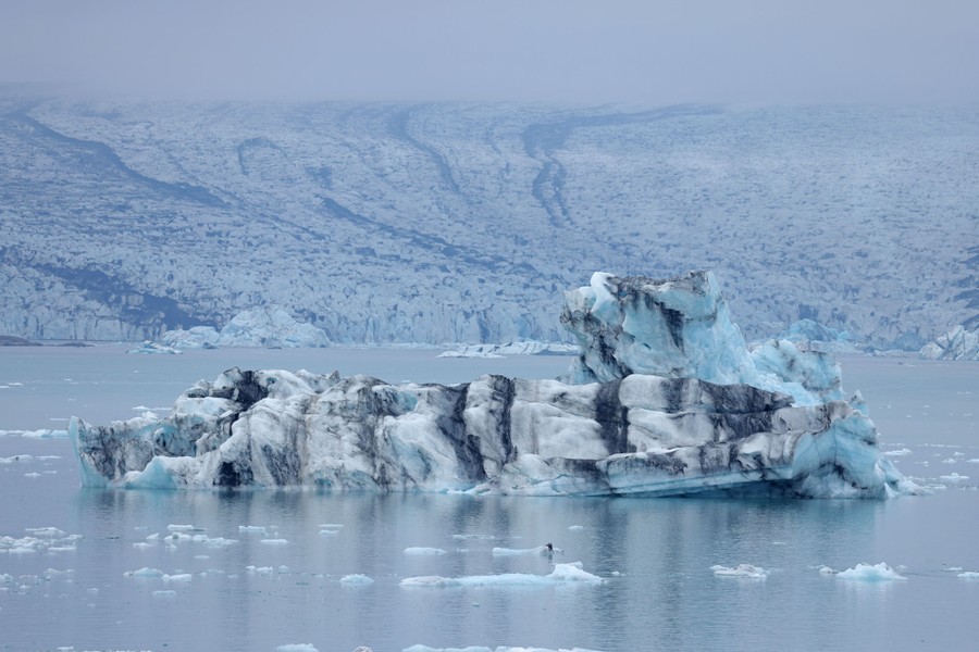 An iceberg striped with rocks floats in a lake in front of a large glacier.