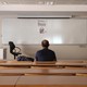 The back of a man sitting in an empty classroom, facing a blank dry-erase board