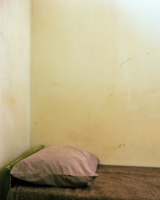 A twin bed in the corner of a room, with a thin brown blanket and a pink pillowcase, against a stained wall