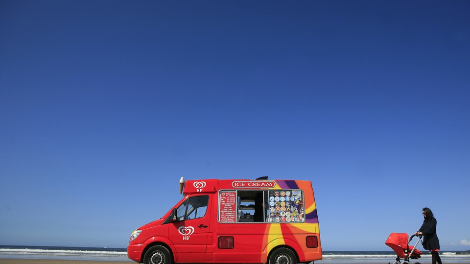 A woman pushing a red pram toward a red ice-cream truck on a beach.