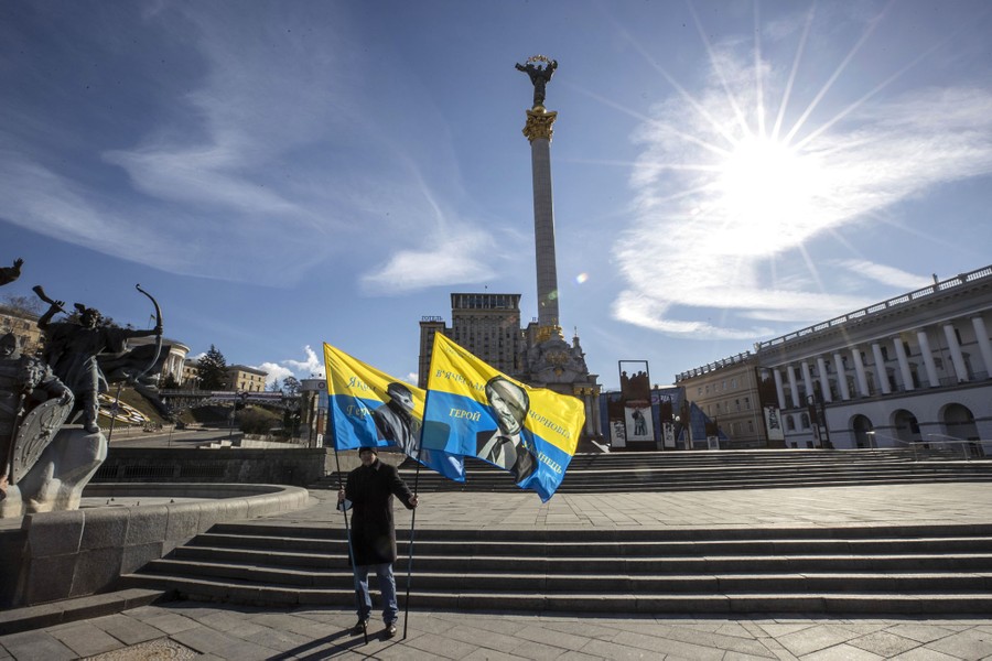 A man stands in a deserted city square holding two Ukrainian flags.