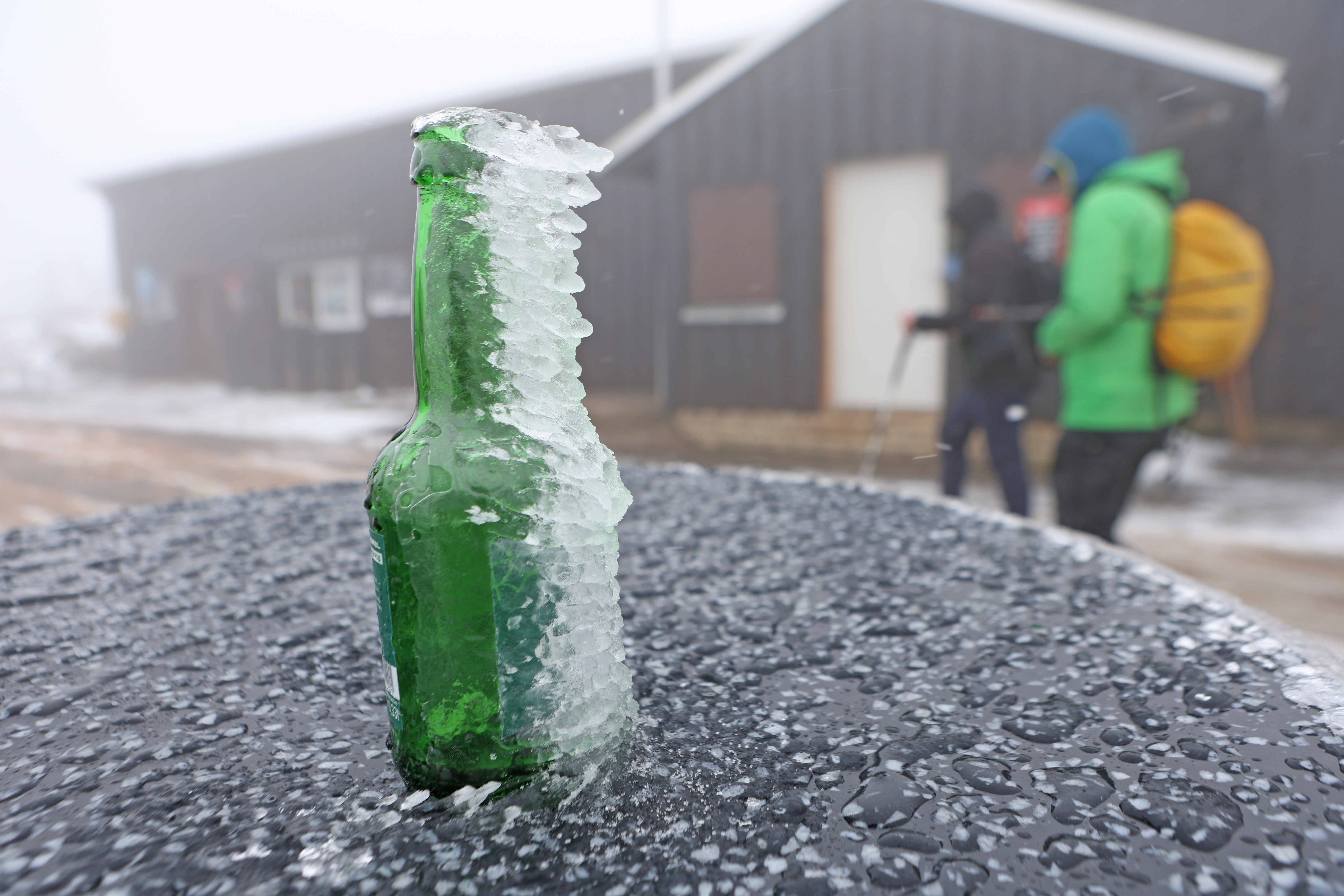 People walk past a green glass bottle covered in wind-blown ice.