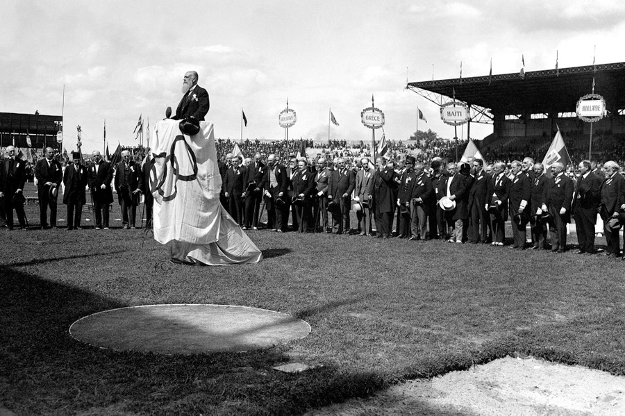 A person stands at a podium draped with an Olympic flag, addressing an audience, while surrounded by men wearing suits, and rows of athletes.