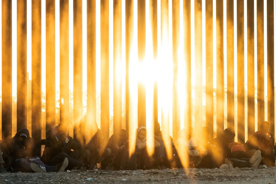 People sit, leaning against a tall border fence, as sunlight shines through slats in the fence.