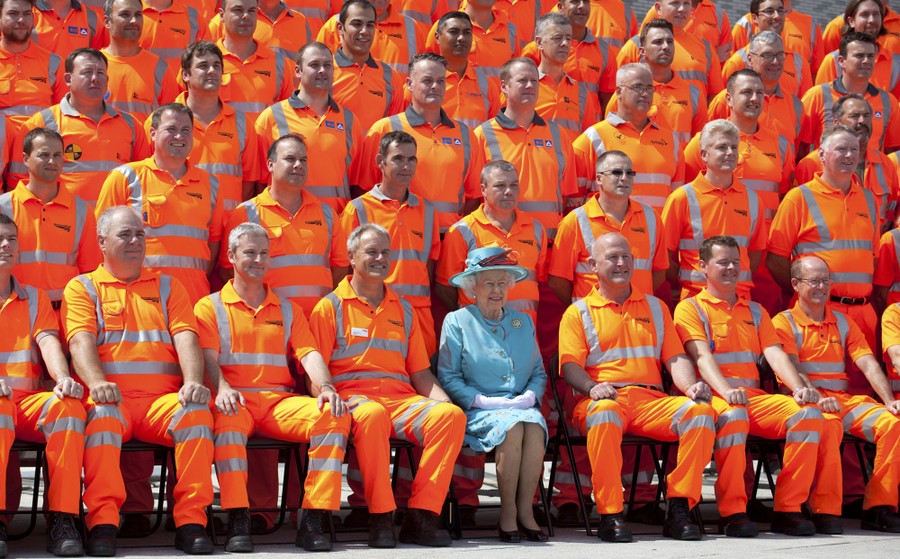 The Queen, in a blue outfit, sits among rows of workers all wearing bright-orange uniforms.