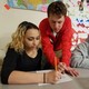 A teacher leans over two students, one of whom is holding a pencil and writing on a worksheet. 