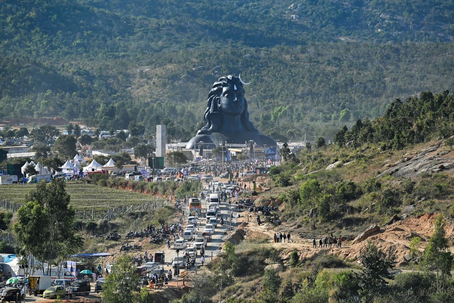 Cars and people crowd around a very tall bust depicting a Hindu god.