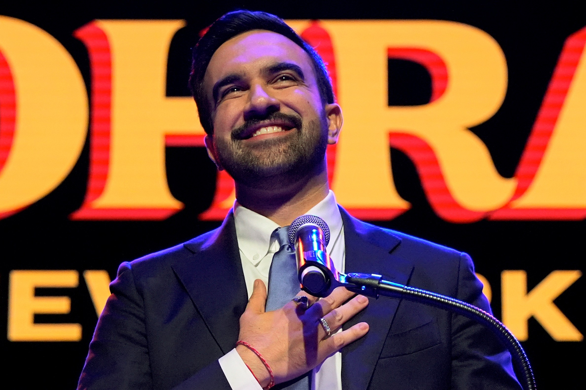New York City mayor-elect Zohran Mamdani stands at a podium, hand on his chest, smiling.