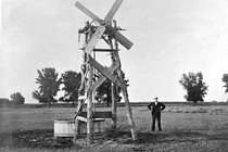 A man stands in a pasture next to a homemade windmill beside a watering trough.