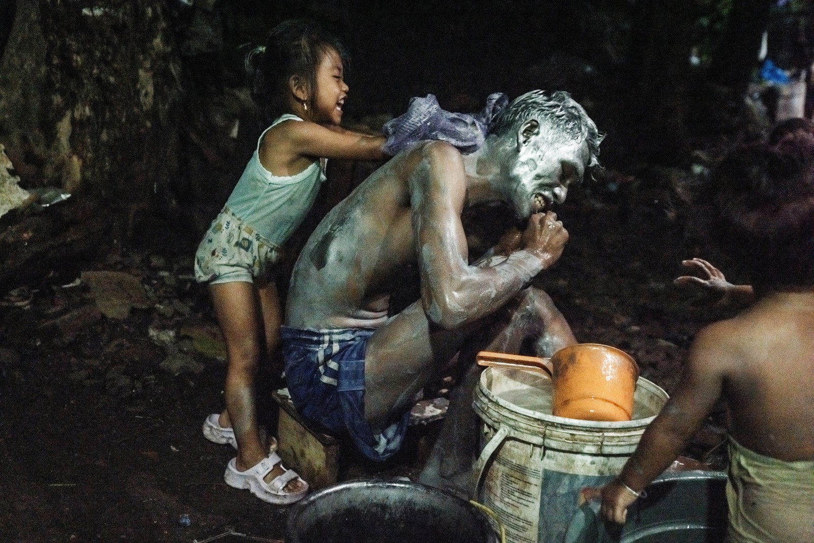 A man kneels down while a young girl playfully scrubs his back with soap and water.