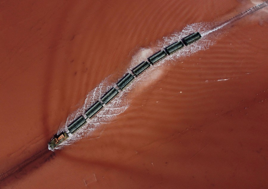 An overhead view of a small train moving on tracks through shallow water, creating a wake