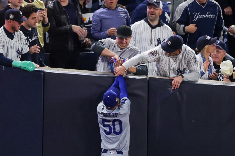 Two baseball fans lean over a wall while pulling a baseball from the glove of a player during a game.