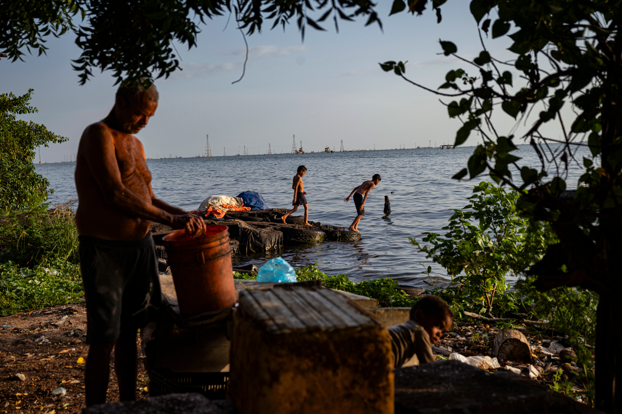 two children play along the shore of Lake Maracaibo in the background, while in the foreground, a man holds an orange bucket