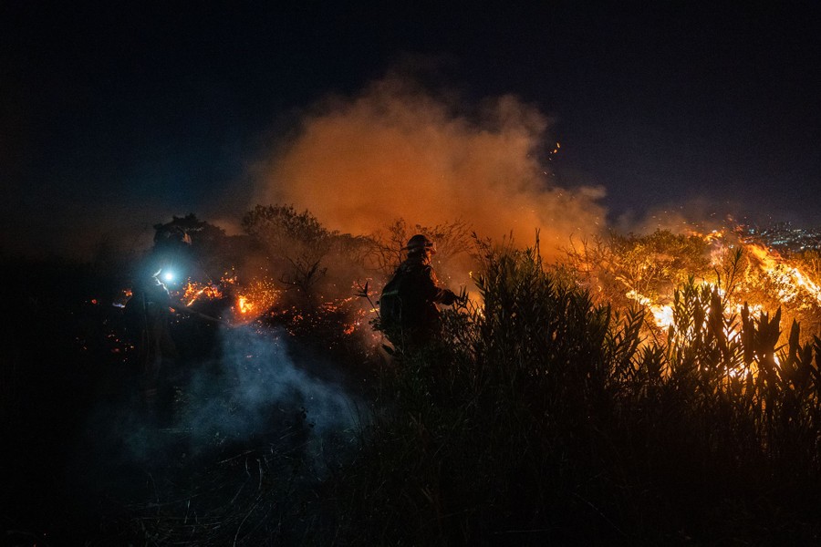 A firefighter walks near a forest fire that burns on a hillside.