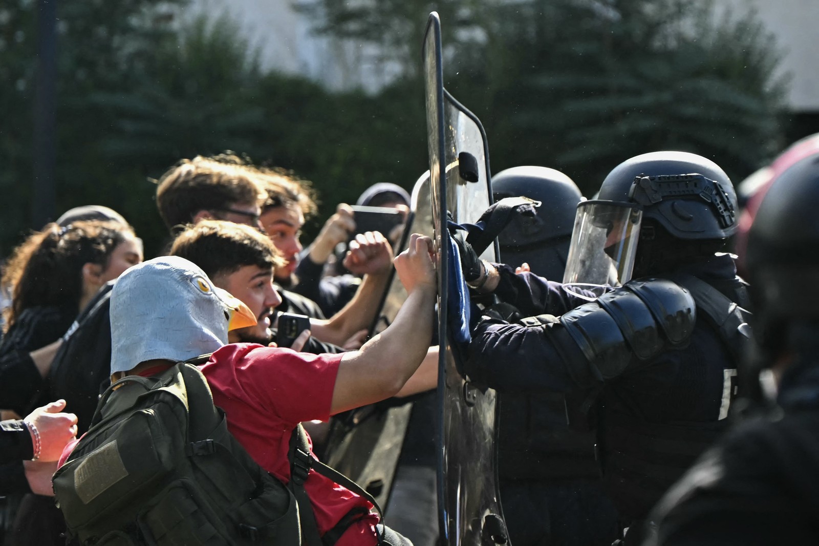 Student protesters, including one wearing a pigeon mask, push on the shields of several riot police.