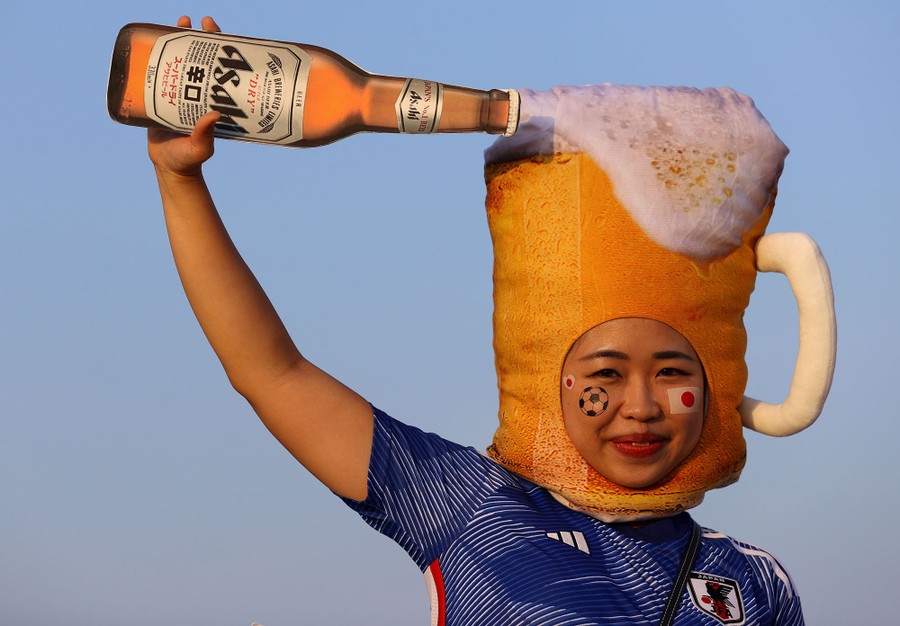 A soccer fan wears a costume hat shaped like a mug of beer, and pretends to pour a large bottle of beer into it.