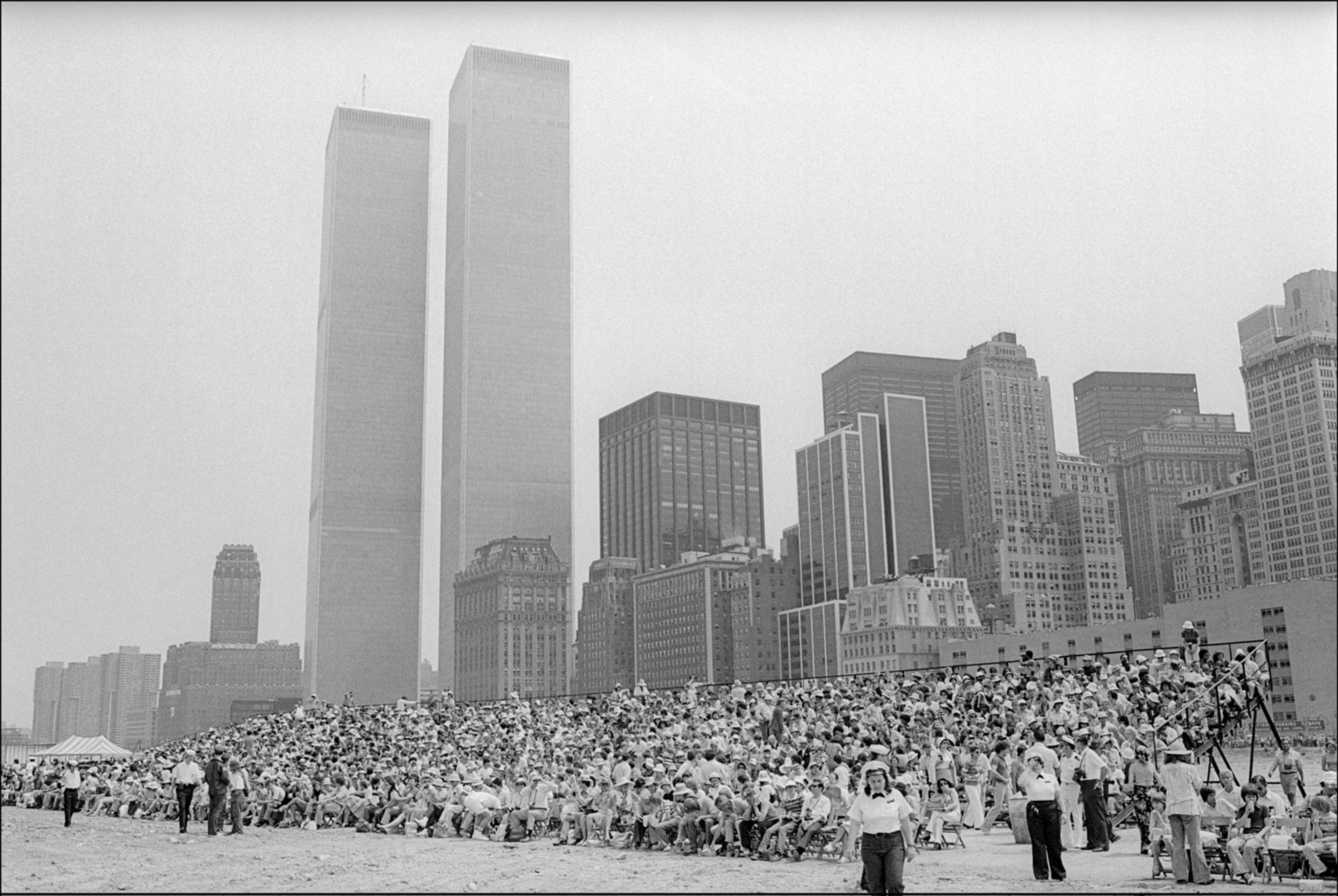 A large crowd sits on bleachers on a shoreline in front of New York City buildings, including the Twin Towers, in 1976.