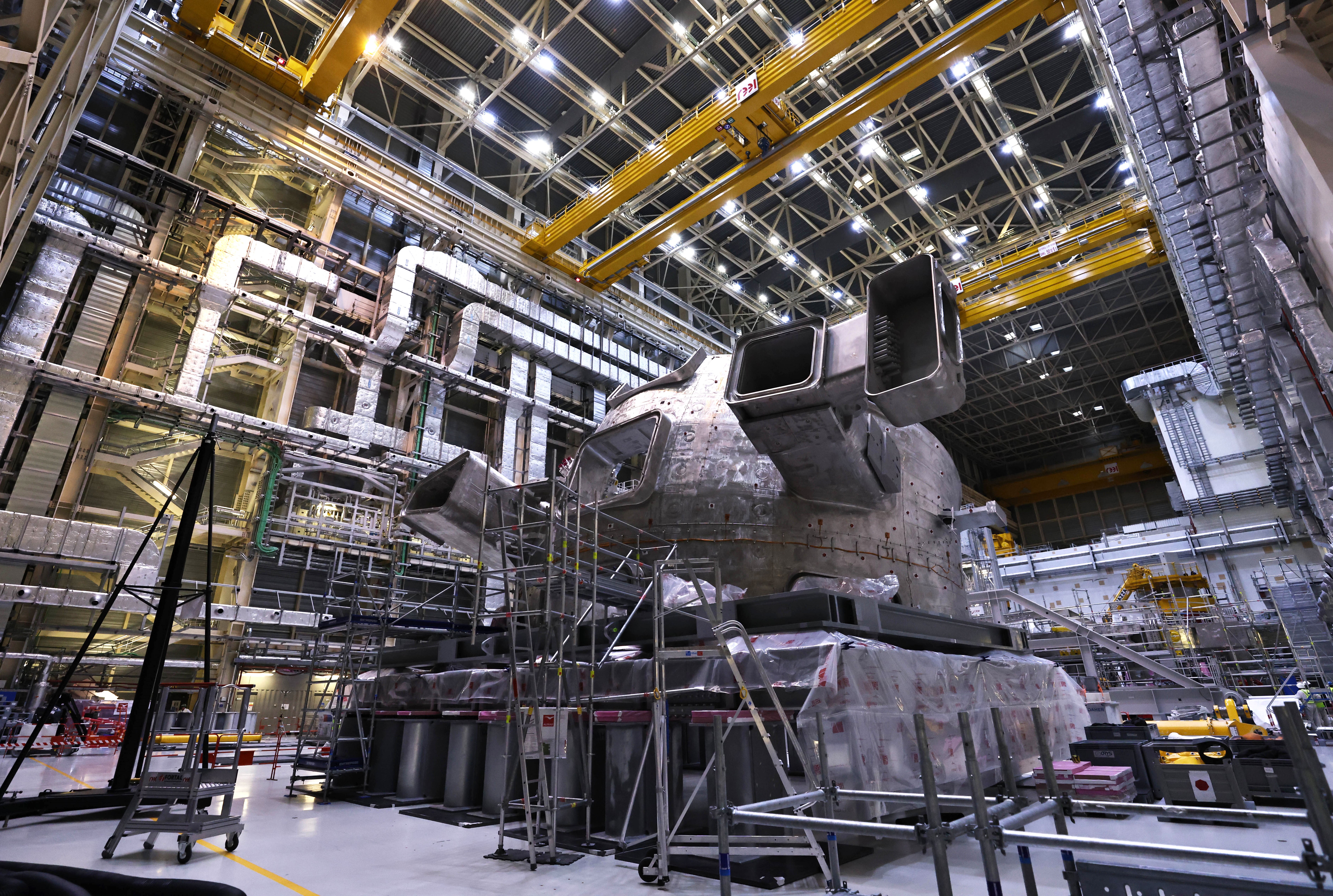 A large metal component of a fusion reactor lies on its side in a huge building, beneath gantry cranes.