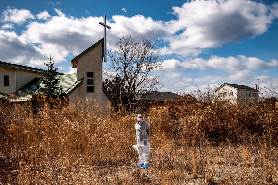 A man in protective gear stands in tall weeds outside an abandoned church building.