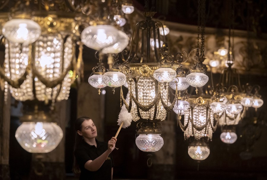 A person uses a duster to clean one of several ornate chandeliers.