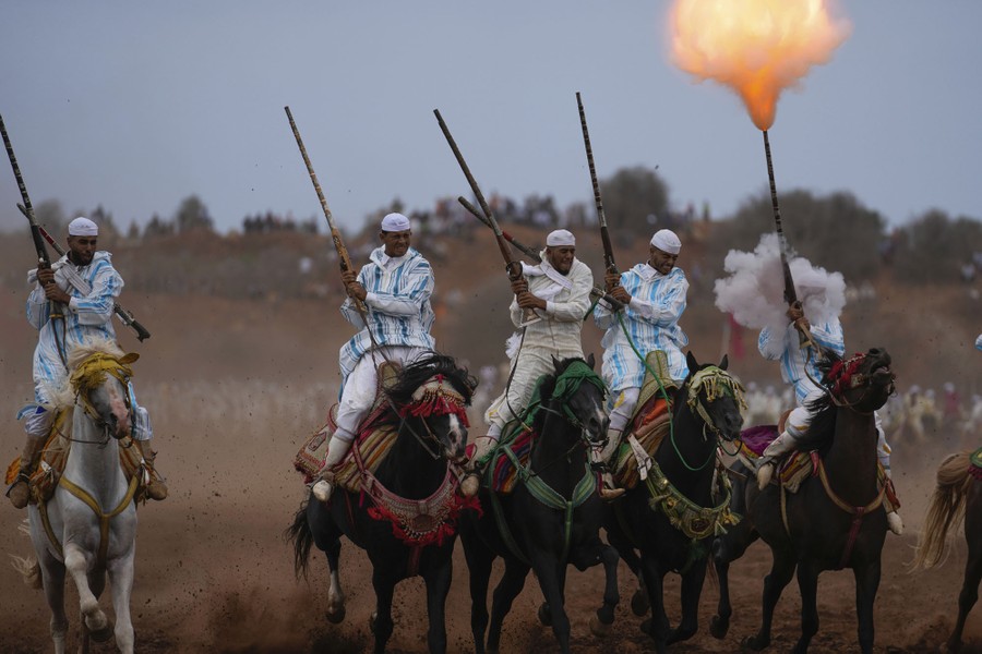 A group of five or six men ride on horseback, carrying rifles. One of them has fired his rifle, creating a puff of smoke by his head, and a small fireball at the end of the muzzle.