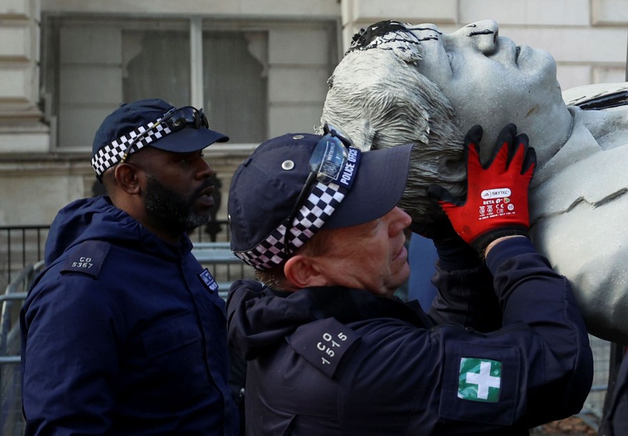 Two police officers load an oil-stained statue of Boris Johnson into a van.