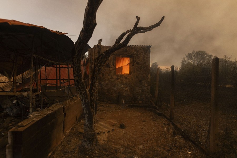 A burned tree and farm structures. An orange glow is seen through the open window of one building.