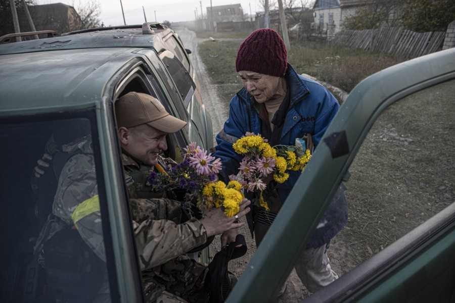 On the side of a road, a person hands flowers to a soldier who is sitting in a truck.