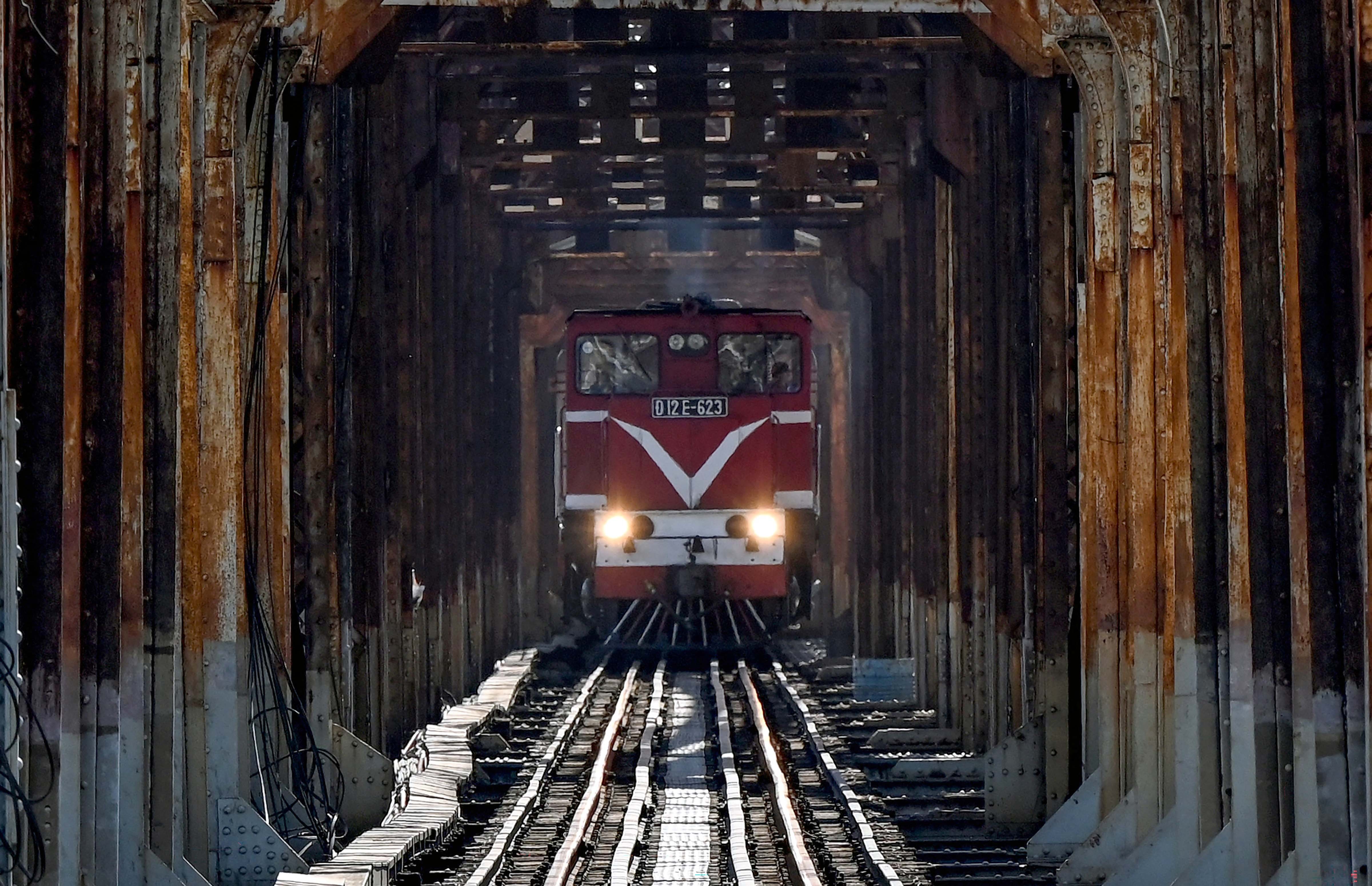A train travels across a rusting steel bridge, seen head-on at a distance.