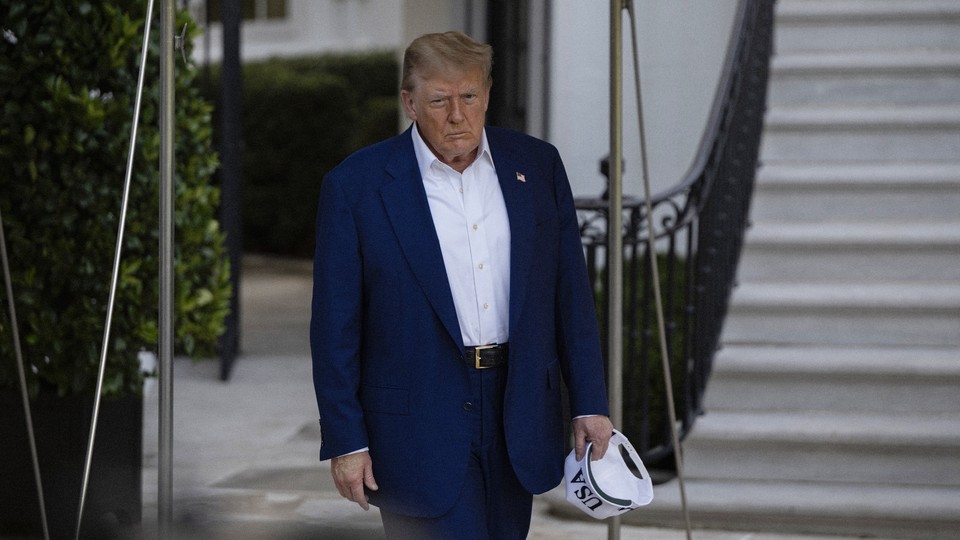 Donald Trump walking, looking displeased, carrying in one hand a hat that reads "USA"