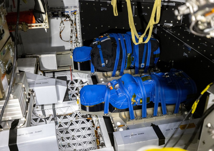 A view of two manikins wrapped in blue cloth and straps, attached to the floor of a spacecraft.