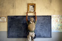 A U.S. Marine, photographed from behind, raises his hands to grip the frame of a portrait of Saddam Hussein hanging above a blackboard.