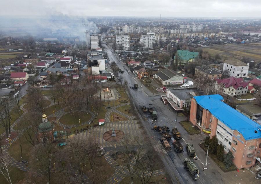 An aerial view of destroyed Russian military vehicles left in a street. Smoke rises from a damaged building in the distance.
