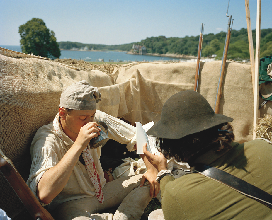 photo of two reenactors dressed as American soldiers resting, one taking a drink of water and the other reading, during a pause in the reenactment, with the shore in the background