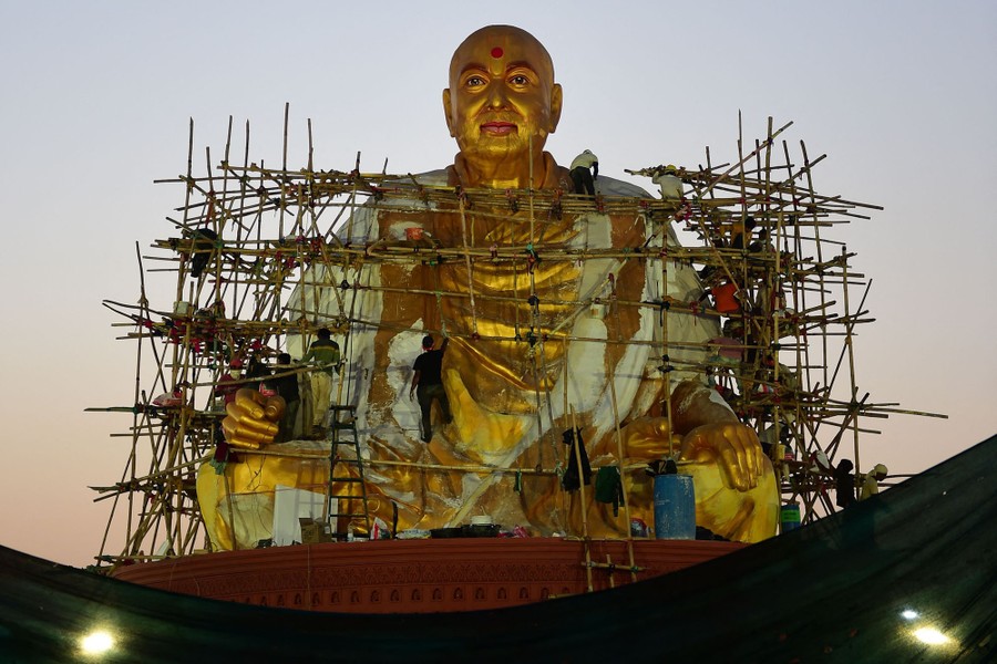 Workers climb scaffolding to work on a large statue of a seated guru.