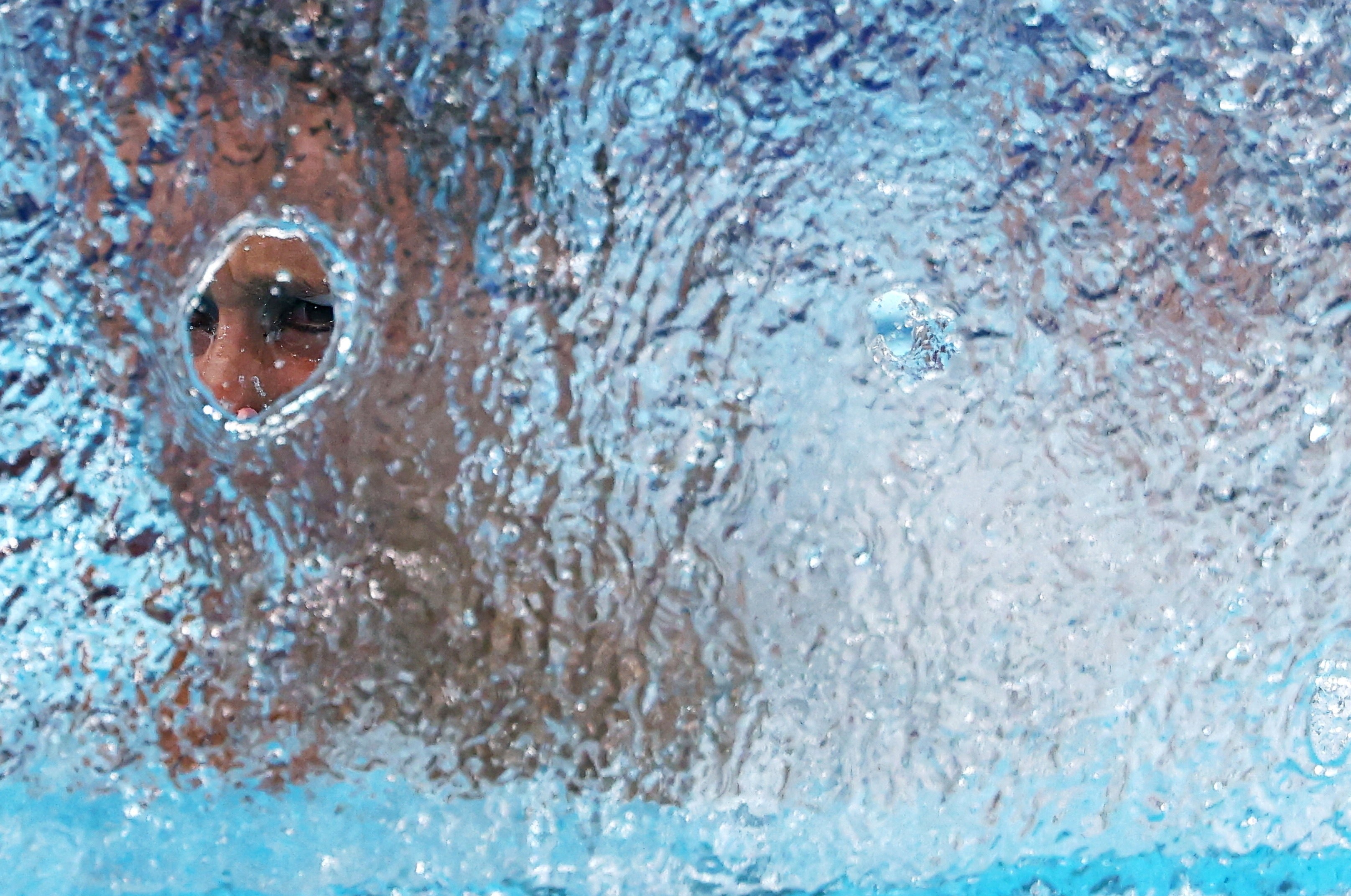 A swimmer performs during a competition, seen through a small hole in a splash of water.