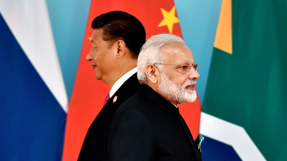 Xi Jinping and Narendra Modi stand next to each other but face in opposite directions in front of flags.